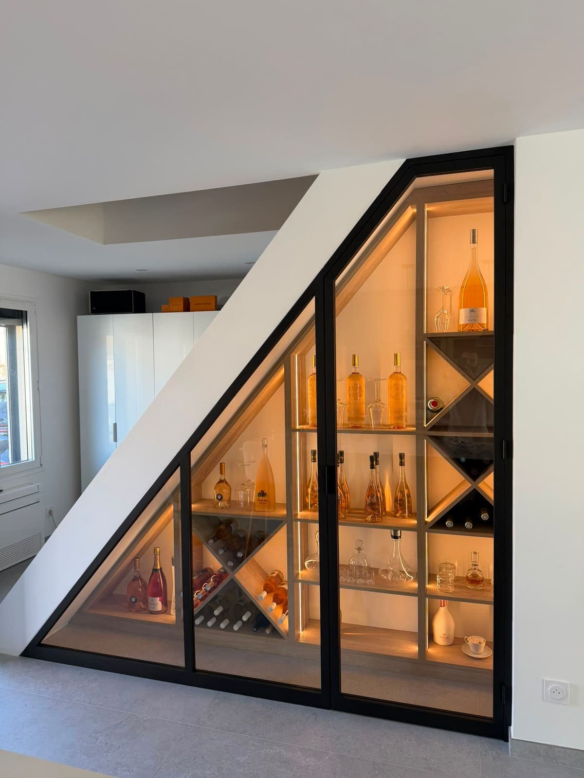 Cave à vin vitrée aménagée sous un escalier, verrière noire et niches éclairées
